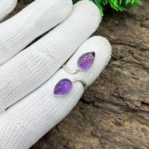 Könnte beinhalten: Ein Silberring mit zwei tropfenförmigen Amethyst-Edelsteinen. Das Band des Rings umschließt den Finger, wobei die violetten Steine in einer Zargenfassung gefasst sind. Der Ring wird auf einem weißen Handschuh vor einem natürlichen Holzgrund gezeigt.