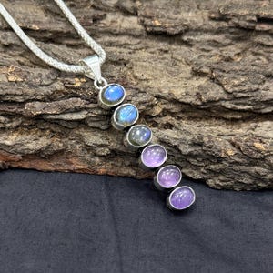 May include: A silver pendant necklace featuring a series of oval-shaped gemstones in shades of blue and purple. The gemstones are set in individual silver bezels and arranged in a vertical line. The necklace chain is a silver woven style.
