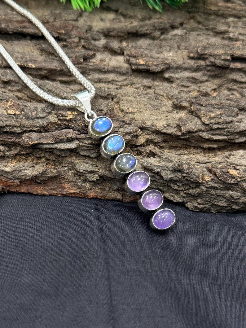 May include: A silver pendant necklace featuring a series of oval-shaped gemstones in shades of blue and purple. The gemstones are set in individual silver bezels and arranged in a vertical line. The necklace chain is a silver woven style.