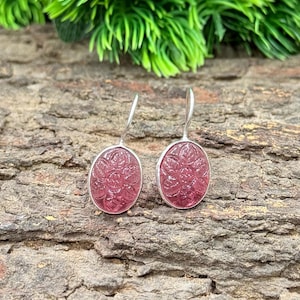 May include: A pair of silver drop earrings. Each earring has an oval, pink gemstone with a floral design. The gemstones are set in a silver bezel and have a polished finish. The earrings are displayed on a textured, brown surface.