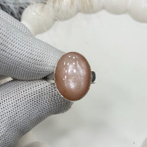 以下が含まれることがあります： 大きな楕円形の桃色の宝石をあしらったシルバーリング。 宝石は滑らかで磨かれた表面を持ち、シンプルなシルバーバンドにセットされています。 リングは白い背景に表示されています。