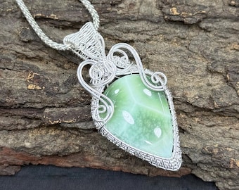 Puede incluir: Un colgante artesanal con una piedra preciosa verde claro, encerrada en un alambre de plata. El colgante cuelga de una delicada cadena de plata. La piedra preciosa tiene una superficie lisa y pulida y tiene forma de lágrima.