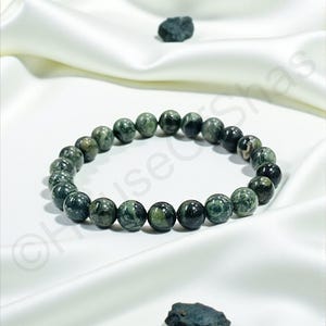 May include: A dark green beaded bracelet with a marbled pattern, resting on a white satin surface. Two small, dark green stones are also present. The bracelet is made of round beads.