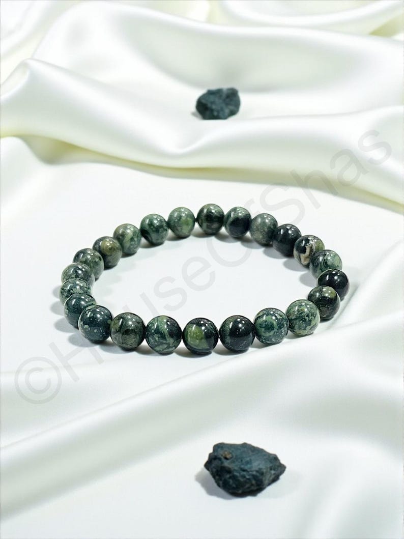 May include: A dark green beaded bracelet with a marbled pattern, resting on a white satin surface. Two small, dark green stones are also present. The bracelet is made of round beads.