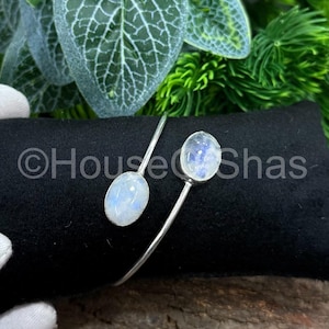 Op de afbeelding: Een zilverkleurige armband met twee ovale maansteen cabochons. De armband heeft een minimalistisch ontwerp met een slanke, gebogen band. De maanstenen vertonen een zachte, iriserende gloed.