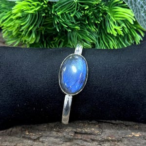 May include: A silver bracelet featuring an oval, iridescent blue gemstone. The gemstone is set in a silver bezel and mounted on a simple silver band. The bracelet is displayed on a black surface with green foliage in the background.