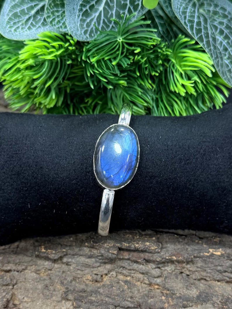 May include: A silver bracelet featuring an oval, iridescent blue gemstone. The gemstone is set in a silver bezel and mounted on a simple silver band. The bracelet is displayed on a black surface with green foliage in the background.