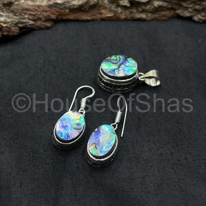 May include: Abalone shell jewelry set featuring a pendant and a pair of earrings. The pendant is round, and the earrings are oval-shaped, all with a silver-colored metal setting. The abalone shells display iridescent blues, greens, and purples.