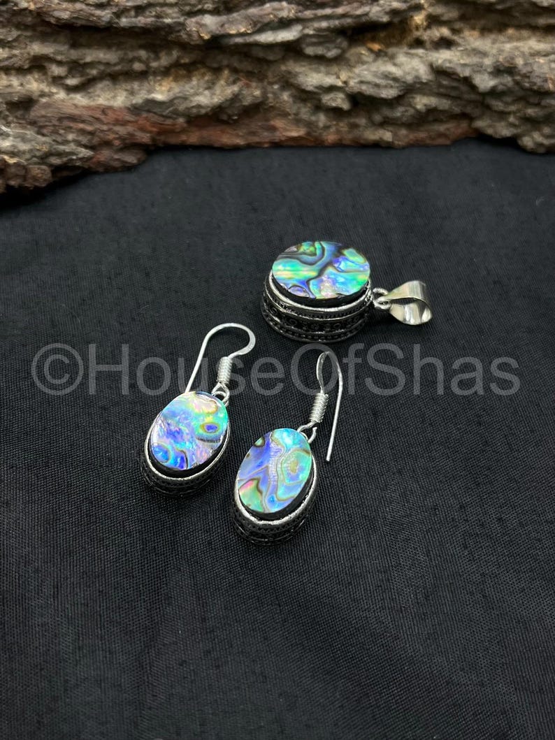 May include: Abalone shell jewelry set featuring a pendant and a pair of earrings. The pendant is round, and the earrings are oval-shaped, all with a silver-colored metal setting. The abalone shells display iridescent blues, greens, and purples.