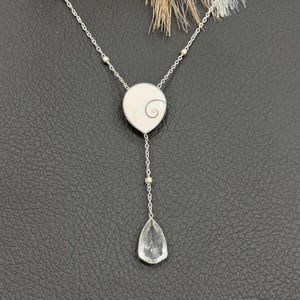May include: A silver necklace with a pendant featuring a white circular design with a spiral and a clear, teardrop-shaped crystal. The necklace has a delicate chain with small silver beads.