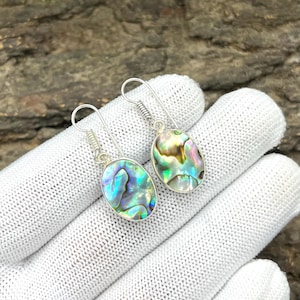 Könnte beinhalten: Ein Paar ovale Ohrringe aus Abalone-Muscheln mit schillernden Blau-, Grün- und Goldtönen. Die Ohrringe haben eine silberfarbene Metalleinfassung und ein einfaches Haken-Design. Die Ohrringe werden auf einer weißen Oberfläche präsentiert.