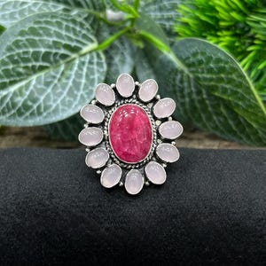 May include: A silver ring featuring a large, oval, deep pink gemstone at the center, surrounded by a halo of smaller, light pink oval stones. The ring is set against a backdrop of green foliage and a black surface.