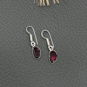 May include: A pair of drop earrings featuring small, irregular, deep red garnet stones set in silver-toned metal. Each earring has a simple silver hook wire with a coiled detail at the top, suspending the polished, dark red gemstone pendant.