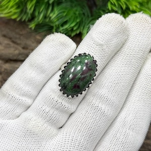 Puede incluir: Un anillo de plata con una piedra preciosa ovalada verde y roja. La piedra preciosa está engastada en un bisel festoneado. El anillo se muestra sobre un guante blanco, con follaje verde en el fondo.