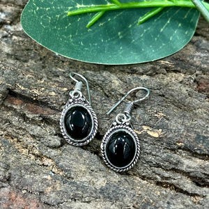 May include: A pair of silver-coloured earrings featuring oval, black gemstones. The earrings have a decorative rope-like border around the black stone and a simple hook closure. The earrings are displayed on a textured, brown surface.