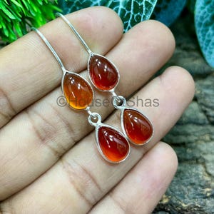 Può includere: Un paio di orecchini pendenti in argento con pietre preziose a forma di goccia, rosso-arancio. Gli orecchini hanno un design semplice ed elegante con una finitura lucida. Le pietre sono incastonate in argento.