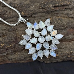May include: A silver pendant necklace featuring a large, starburst-shaped pendant. The pendant is adorned with numerous pear-shaped, iridescent moonstone cabochons. The necklace chain is a delicate, textured silver. The background is a dark, textured surface.