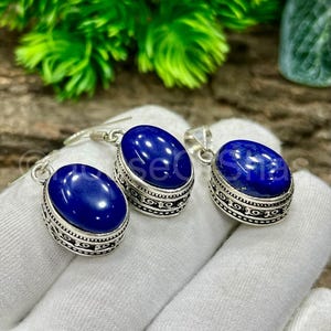 Puede incluir: Un conjunto de joyas con un par de pendientes ovalados y dos colgantes. Cada pieza tiene una piedra preciosa azul oscuro pulida engastada en un marco decorativo de metal plateado. El conjunto se muestra sobre una superficie blanca.