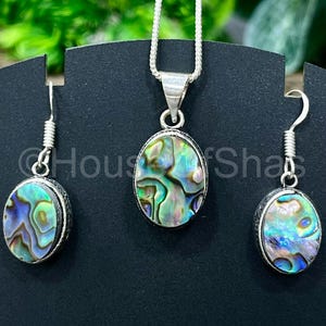 May include: Abalone shell jewelry set featuring a pendant necklace and matching earrings. The oval-shaped abalone shells display iridescent blues, greens, and purples, set in silver-toned metal. The earrings have a hook closure. The necklace has a silver chain.