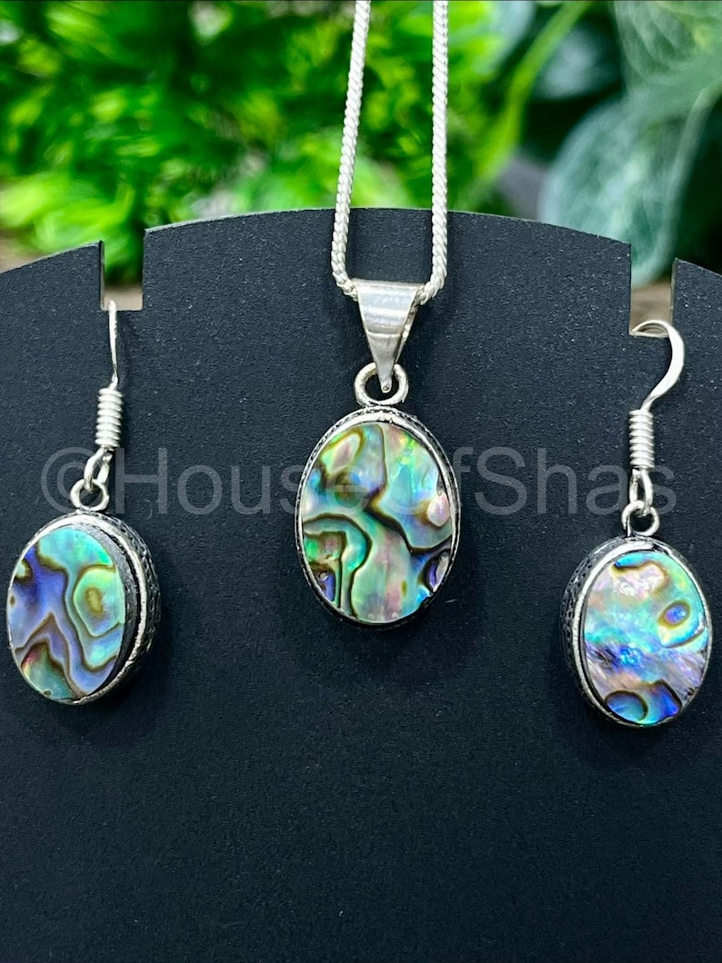 May include: Abalone shell jewelry set featuring a pendant necklace and matching earrings. The oval-shaped abalone shells display iridescent blues, greens, and purples, set in silver-toned metal. The earrings have a hook closure. The necklace has a silver chain.
