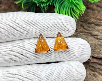 May include: A pair of triangular amber earrings with a silver border. The earrings are set against a white knitted background, with green foliage and wood in the background. The amber has a warm, golden color.