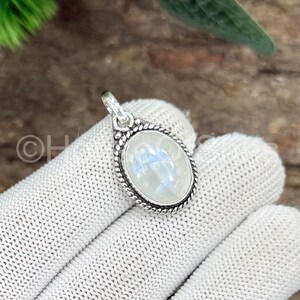May include: Oval-shaped moonstone pendant with a silver-colored, textured border. The iridescent, white moonstone is set in a detailed, decorative frame. The pendant is suspended from a small loop, ready to be added to a necklace.
