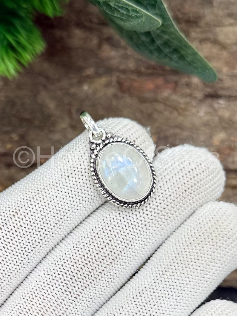 May include: Oval-shaped moonstone pendant with a silver-colored, textured border. The iridescent, white moonstone is set in a detailed, decorative frame. The pendant is suspended from a small loop, ready to be added to a necklace.