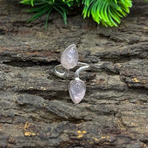 May include: A silver ring featuring two teardrop-shaped rose quartz stones. The stones are a pale pink color and are set in a swirling silver band. The ring is displayed on a textured, dark brown surface with green foliage in the background.