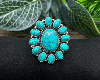 Puede incluir: Anillo plateado con una gran piedra turquesa ovalada en el centro, rodeada de piedras turquesas más pequeñas. El anillo tiene un engaste detallado y ornamentado, y las piedras turquesas tienen un patrón de matriz distintivo.