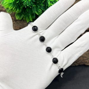 Peut inclure: Un bracelet en argent orné d'une série de grosses pierres noires rondes et polies. Le bracelet est présenté sur une surface blanche, avec une plante verte en arrière-plan. Il possède une chaîne délicate et un charm décoratif.