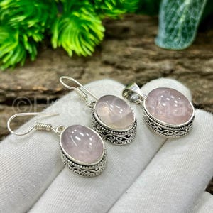 May include: A jewelry set featuring a pair of earrings and a pendant. Each piece has a light pink, oval-shaped stone set in a silver-colored, ornate frame. The earrings have a hook closure.