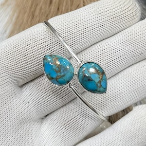 May include: A silver-toned bracelet featuring two teardrop-shaped turquoise stones with copper matrix. The stones are set in a minimalist design, creating a striking contrast against the silver band. The bracelet is shown on a white surface.