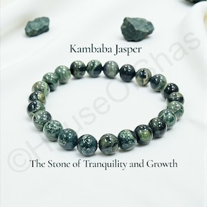 May include: A Kambaba Jasper beaded bracelet with green and black spherical stones. The bracelet is displayed on a white cloth, with the text "Kambaba Jasper" and "The Stone of Tranquility and Growth".
