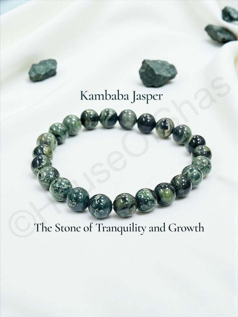 May include: A Kambaba Jasper beaded bracelet with green and black spherical stones. The bracelet is displayed on a white cloth, with the text "Kambaba Jasper" and "The Stone of Tranquility and Growth".