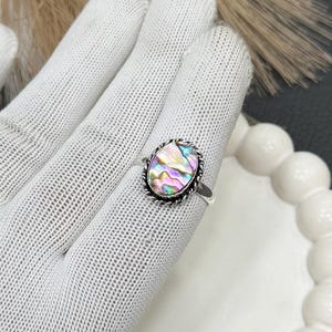 May include: Oval-shaped abalone shell ring with a silver band. The iridescent shell is surrounded by a twisted rope design. The ring is displayed on a white glove.