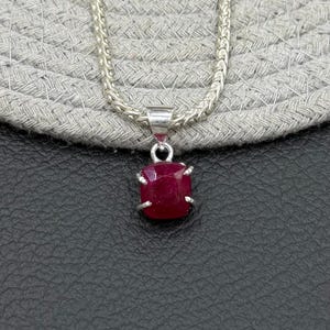 Peut inclure: Un collier à chaîne en argent avec un pendentif rubis rouge foncé de forme carrée, maintenu par quatre griffes en argent. Le pendentif est suspendu à une chaîne en argent métallique à la texture tressée, formant un bijou simple et classique.