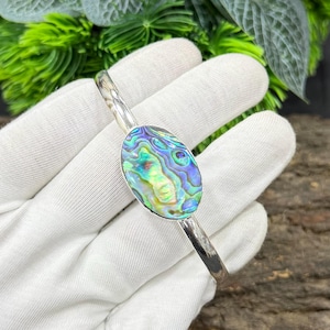 Pode incluir: Uma pulseira de punho prateada com uma pe&ccedil;a central oval de concha de abalone. A concha exibe azuis, verdes e roxos iridescentes. A pulseira &eacute; exibida em um fundo branco com folhagem verde.