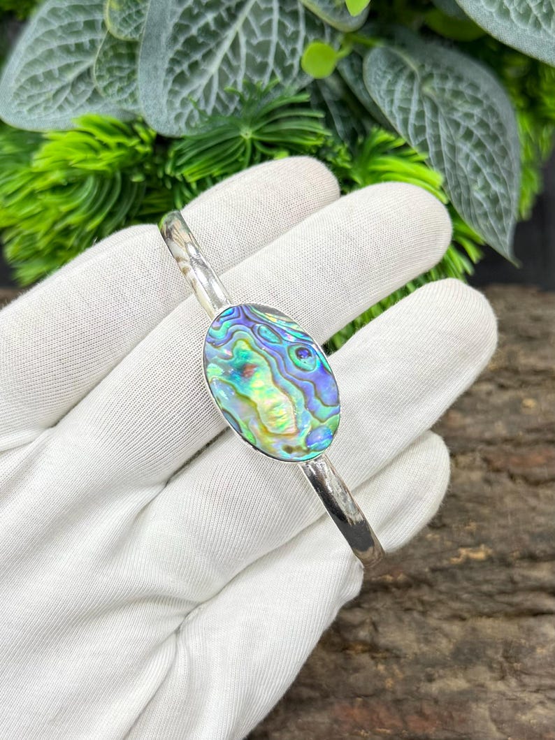 Pode incluir: Uma pulseira de punho prateada com uma pe&ccedil;a central oval de concha de abalone. A concha exibe azuis, verdes e roxos iridescentes. A pulseira &eacute; exibida em um fundo branco com folhagem verde.