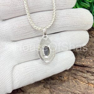 May include: A silver necklace with a teardrop pendant. The pendant has a dark stone set in the center. The chain is a delicate, linked design. The necklace is displayed on a white surface.