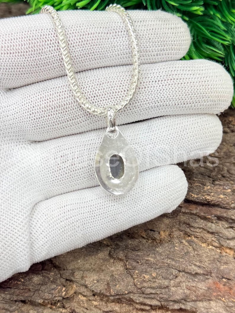 May include: A silver necklace with a teardrop pendant. The pendant has a dark stone set in the center. The chain is a delicate, linked design. The necklace is displayed on a white surface.