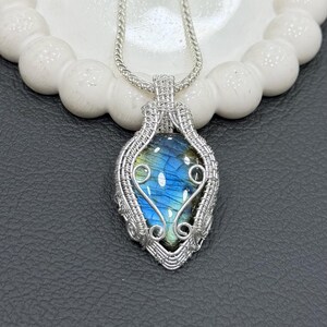 May include: A handcrafted silver pendant necklace featuring a teardrop-shaped labradorite stone. The stone displays iridescent blue and green hues, encased in a detailed wire-wrapped setting. The pendant hangs from a delicate silver chain.
