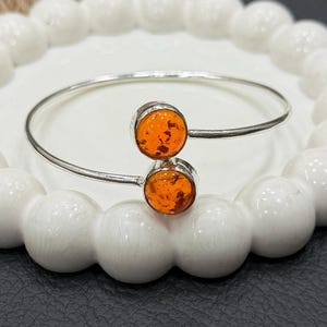 May include: A silver bangle bracelet featuring two round, orange amber stones. The amber stones have a warm, translucent appearance and are set in a silver bezel. The bracelet is displayed on a white, decorative plate.