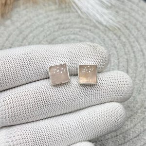 Op de afbeelding: Een paar vierkante oorknopjes met lichtroze, doorschijnende stenen in zilverkleurige metalen zettingen. De oorbellen hebben een glad, gepolijst oppervlak en een eenvoudig, geometrisch ontwerp met een zachte, bleke roze kleur.