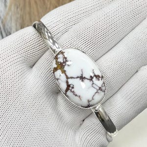 May include: A silver cuff bracelet featuring an oval white stone with brown and black veining. The stone is set in a silver bezel and the bracelet has a simple, polished design. This piece of jewelry is suitable for everyday wear.