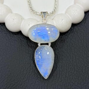 Peut inclure: Un collier pendentif en argent avec deux pierres de lune. La pierre supérieure est ovale et l'inférieure en forme de goutte d'eau. Les deux pierres ont une lueur irisée bleue et blanche. La chaîne du collier est de style corde argentée.