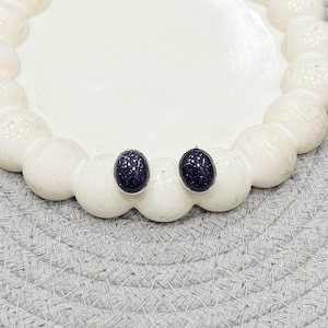 May include: A pair of oval-shaped stud earrings featuring dark blue, glittery goldstone cabochons set in simple silver-toned metal frames. The stones have a deep navy hue with numerous tiny, reflective metallic flecks that mimic a starry night sky.