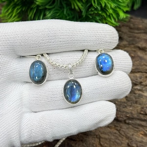May include: A silver necklace and matching earrings set featuring oval-shaped labradorite stones. The stones display a blue and green iridescent sheen. The jewelry is displayed on a white glove against a backdrop of greenery and wood.