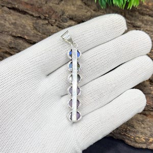 May include: A silver pendant featuring a vertical design with a series of colorful, round gemstones. The gemstones include blue, green, and purple hues. The pendant is held by a white gloved hand.