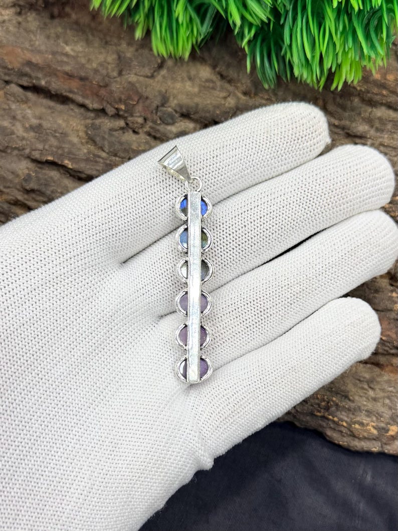 May include: A silver pendant featuring a vertical design with a series of colorful, round gemstones. The gemstones include blue, green, and purple hues. The pendant is held by a white gloved hand.