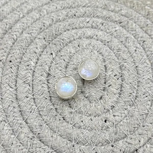 Pode incluir: Um par de brincos redondos. Cada brinco apresenta uma pedra iridescente branca e azul embutida em uma moldura prateada. Os brincos são exibidos em uma superfície cinza tecida.